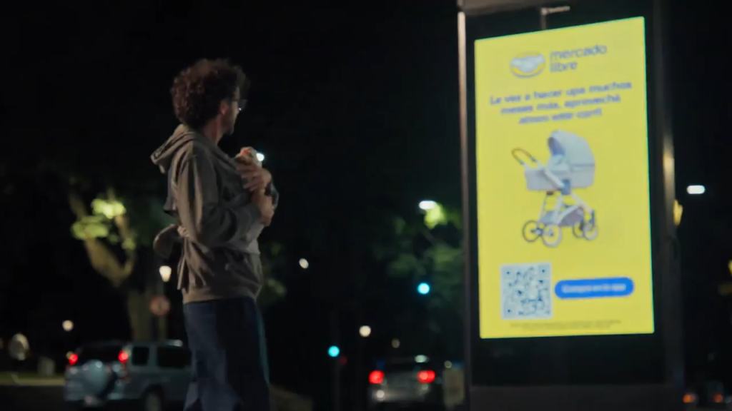 Videoframe da campanha do Mercado Livre. Uma pessoa segura um bebê no colo enquanto observa um painel digital iluminado em um ponto de ônibus durante a noite. Na tela, há um anúncio com a imagem de um carrinho de bebê, QR Code e botão de chamada para ação. Ao fundo, aparecem árvores, postes de iluminação e carros circulando pela via.